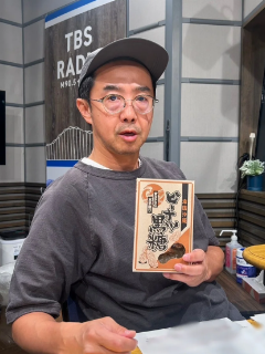 おぎやはぎの矢作兼が帽子を被り、眼鏡をかけ、左手に黒糖を持ちながらカメラに向かって決め顔をしている写真